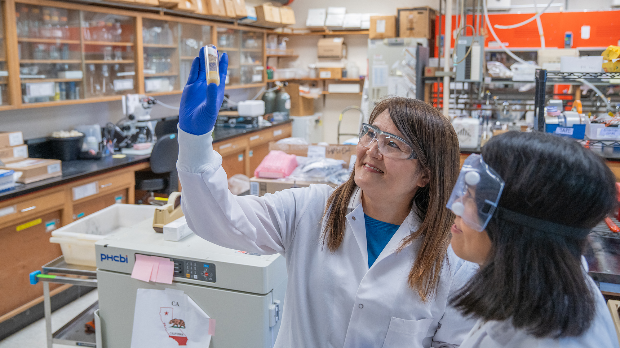 Scientist in lab coat holds a vial up to the light while speaking with a student in safety goggles.