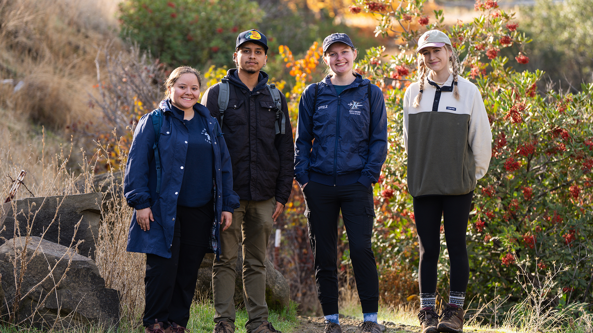 Four people stand side by side outdoors, smiling at the camera in front of a background of dry grass and a bush with red berries; all are dressed in outdoor gear, suggesting participation in a field activity.