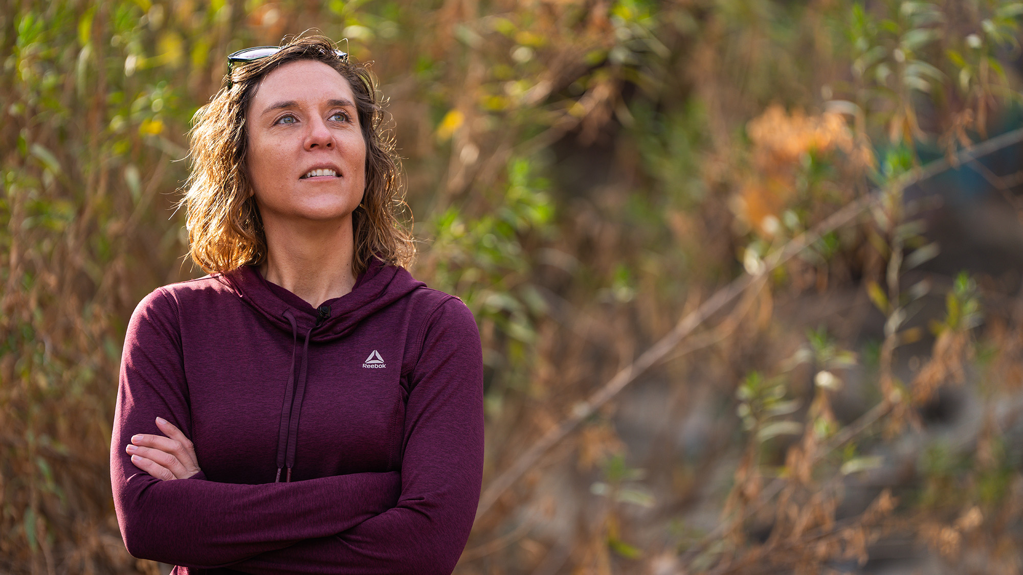 Alt text: Person wearing a maroon hoodie stands outdoors with arms crossed, looking thoughtfully into the distance; background shows dry vegetation and soft sunlight.