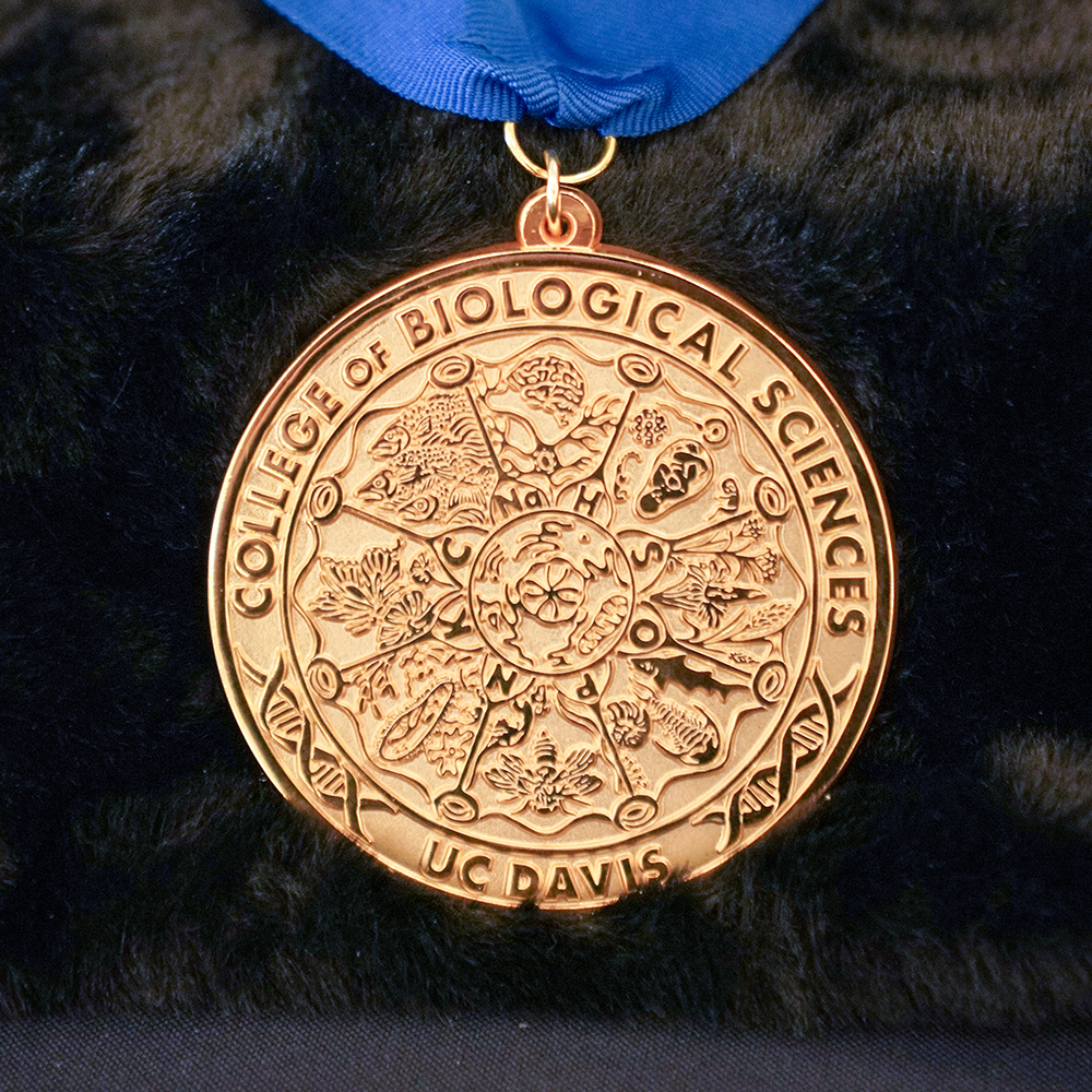 Gold CBS medal with diatom-inspired design and blue ribbon displayed on black fabric.