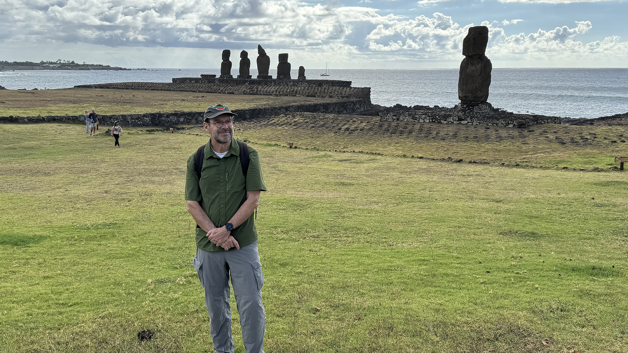 A man in a green shirt and gray pants stands smiling on a grassy field with a backpack, in front of a coastal archaeological site on Easter Island. In the background, a row of moai statues stands on a stone platform near the ocean, with one lone statue off to the right. A few people walk in the distance, and a sailboat is visible on the water under a partly cloudy sky.