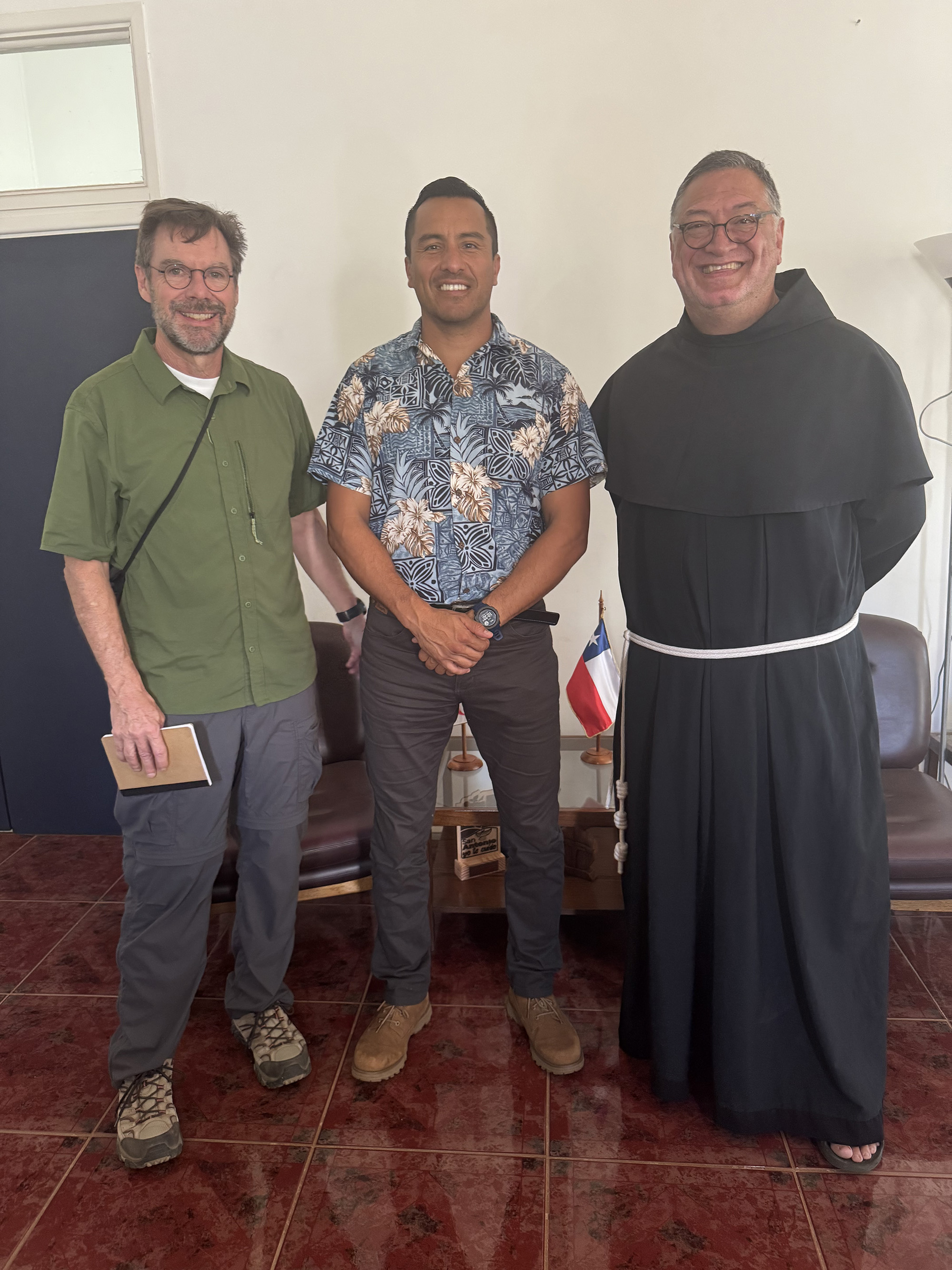 Three men standing indoors and smiling at the camera. The man on the left wears a green shirt and holds a notebook, the man in the middle wears a floral patterned shirt, and the man on the right is dressed in a black robe with a white cord. Behind them are chairs, a small table with miniature flags, and a plain white wall.