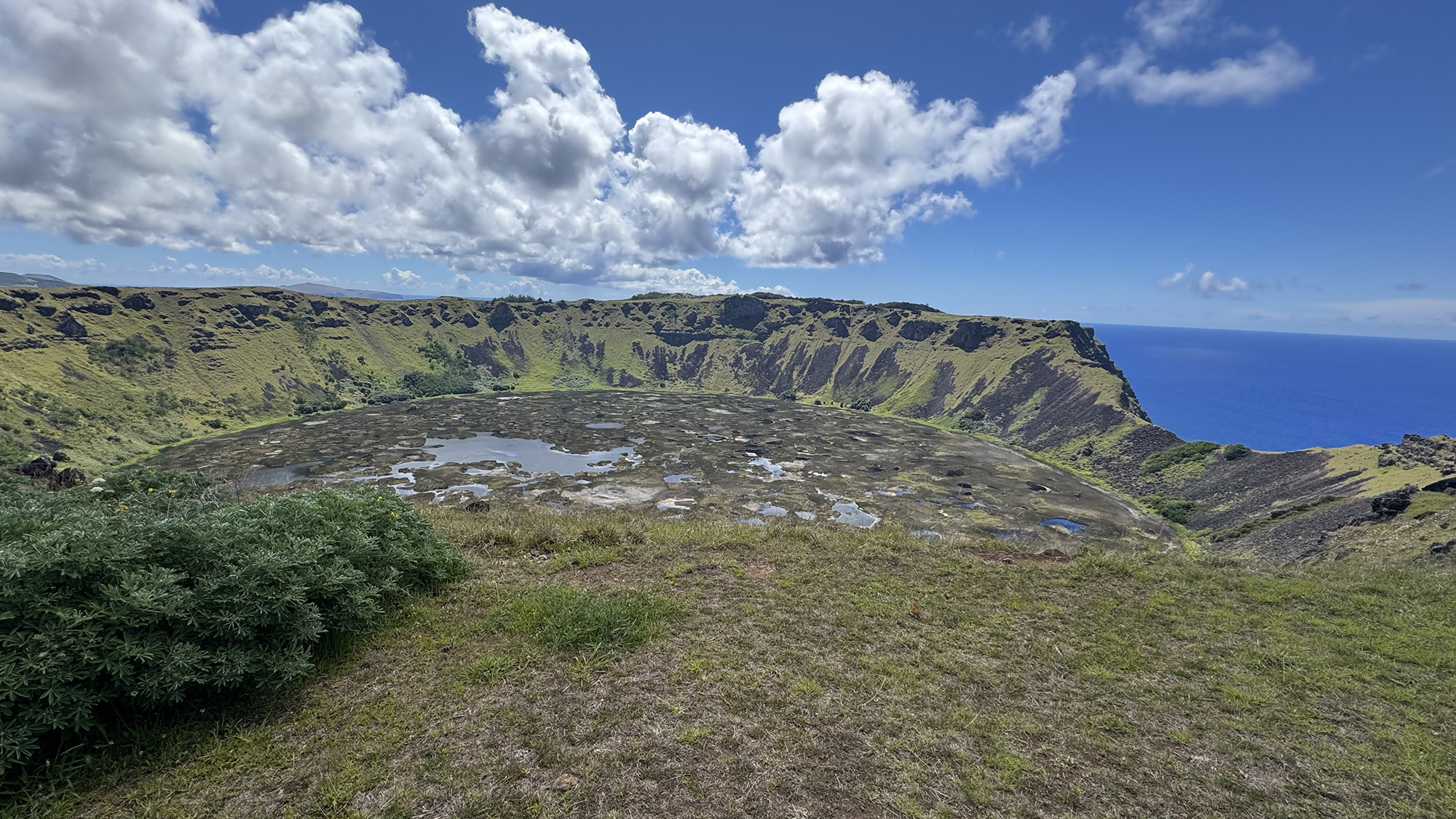 View of a large volcanic crater filled with patches of water and vegetation, surrounded by steep green cliffs. In the background, the crater rim meets a deep blue ocean under a bright sky with scattered clouds. Grassy terrain and shrubs are visible in the foreground.