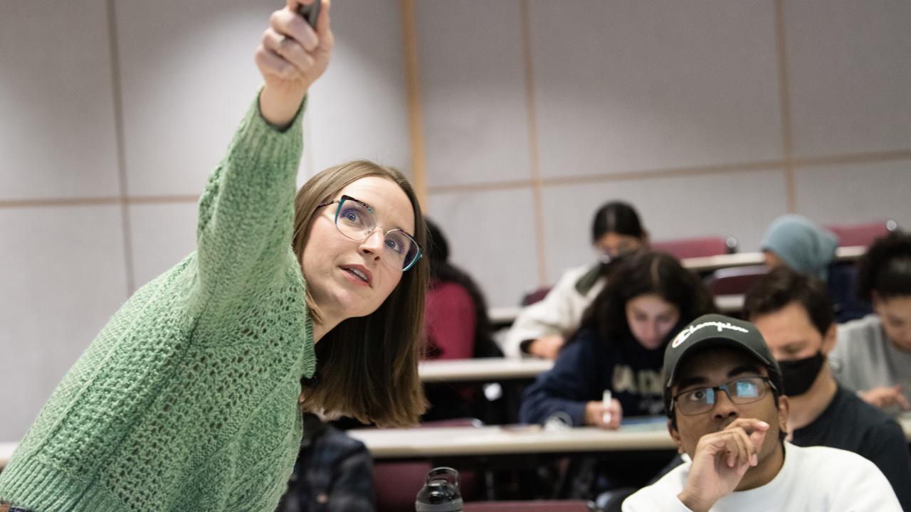 A professor in a green sweater points at a screen while a student in a black cap listens intently; other students take notes in the background.