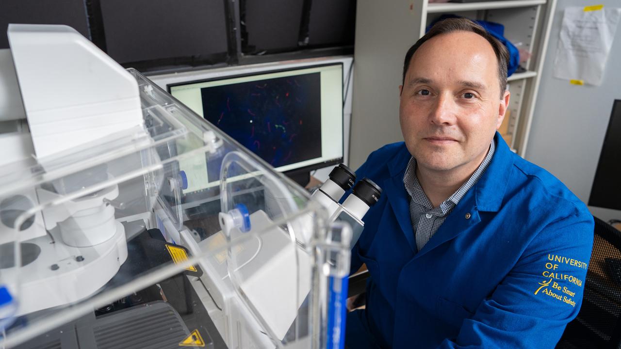 A scientist in a blue University of California lab coat sits beside a microscope and imaging equipment in a research lab. A computer screen behind him displays multicolored microscopic images.