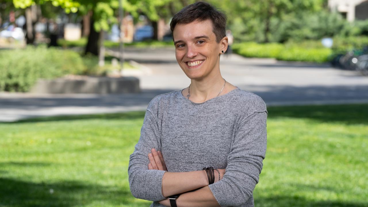A person with short hair and a gray long-sleeve shirt stands smiling with arms crossed on a sunny, tree-lined lawn at UC Davis.