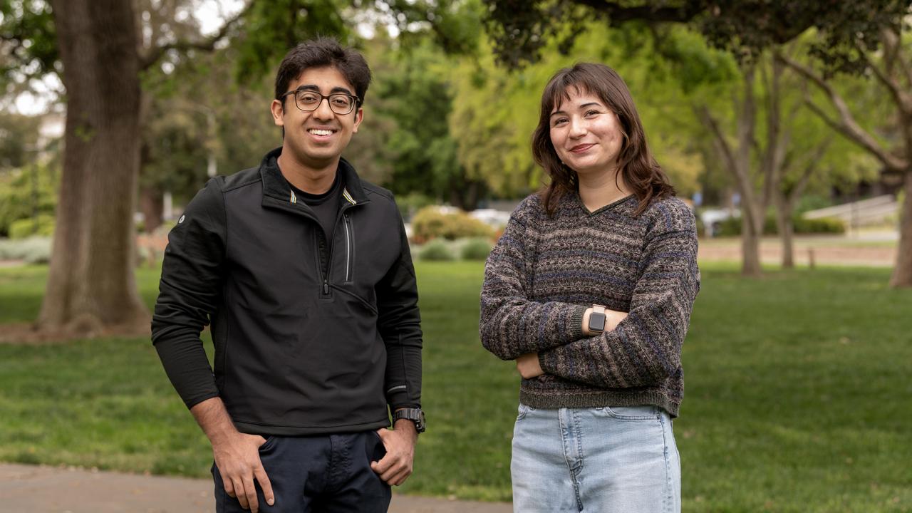 Two students standing outdoors on a green lawn, smiling at the camera. The student on the left wears glasses and a black jacket with hands in pockets, while the student on the right wears a multicolored sweater and light jeans with arms crossed. Trees and pathways are visible in the background.