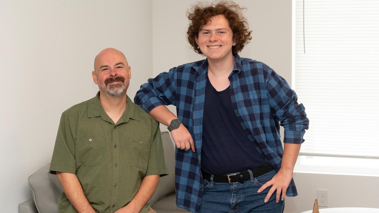 Two people smile in a bright room one seated on a couch and the other standing with an arm resting on the couch back.