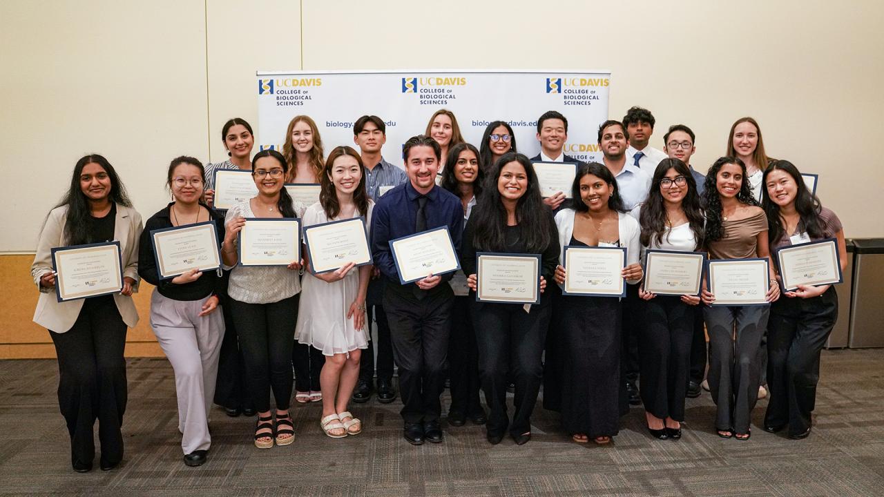 A group of UC Davis students pose together indoors, smiling and holding certificates during an award ceremony hosted by the College of Biological Sciences. A branded backdrop with the UC Davis logo is visible behind them.