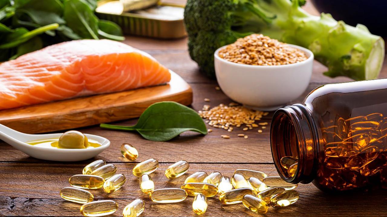A close-up of nutrient-rich foods and supplements on a wooden surface, featuring a raw salmon fillet on a cutting board, a bowl of sesame seeds, fresh spinach leaves, broccoli, and a white spoon containing olive oil and an olive. Nearby, an amber bottle is tipped over, spilling golden omega-3 capsules.