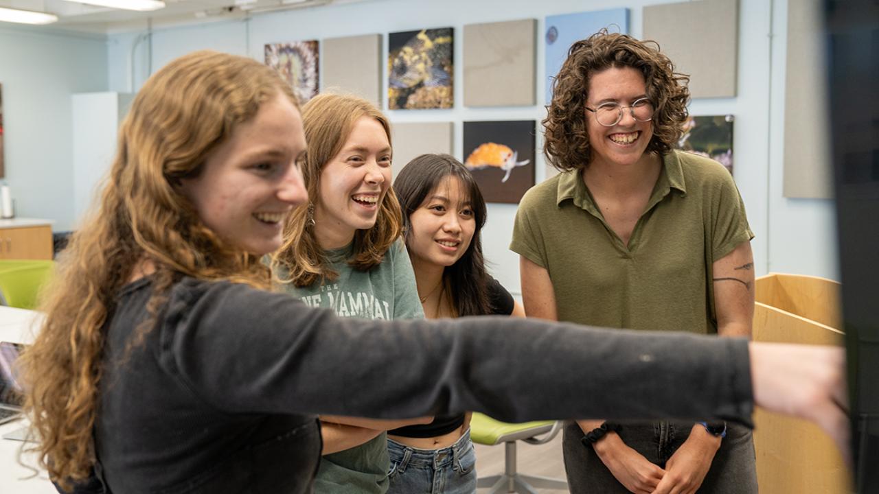 Four students smile and laugh while looking at a screen in a classroom or lab setting. One student in the foreground points at the screen, while the others stand closely together. The room has colorful photos of marine life on the wall in the background.
