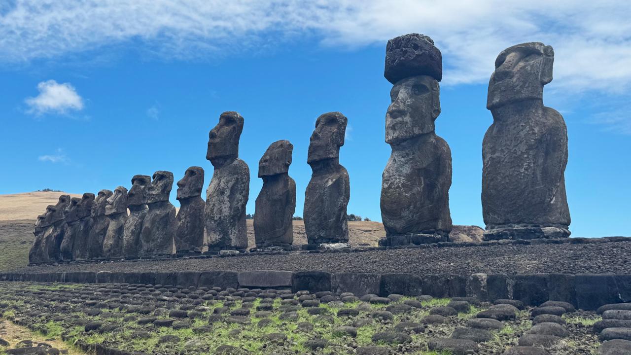 A row of large stone moai statues standing on a stone platform (ahu) at Ahu Tongariki on Easter Island. The statues face inland, with one wearing a topknot-like stone hat (pukao). The background shows grassy hills and a bright blue sky with scattered clouds.