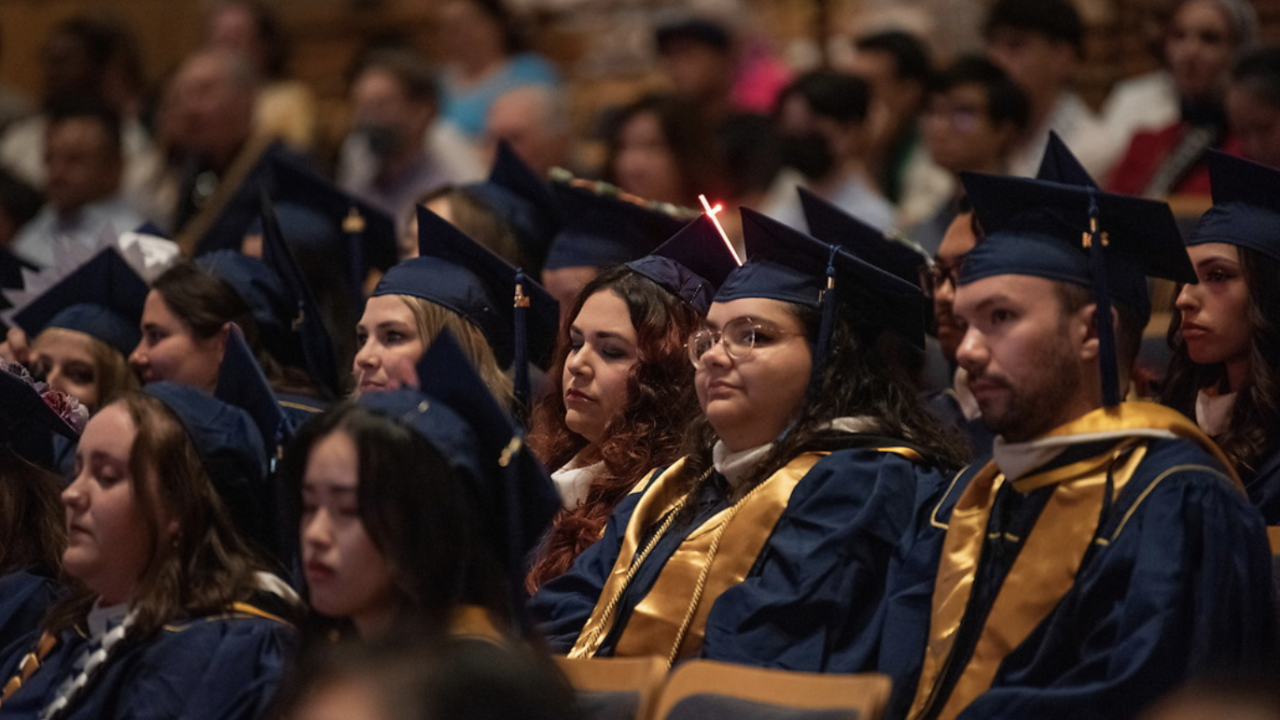 Graduates in blue and gold caps and gowns sit attentively in an auditorium, with one student's cap featuring a glowing red light.