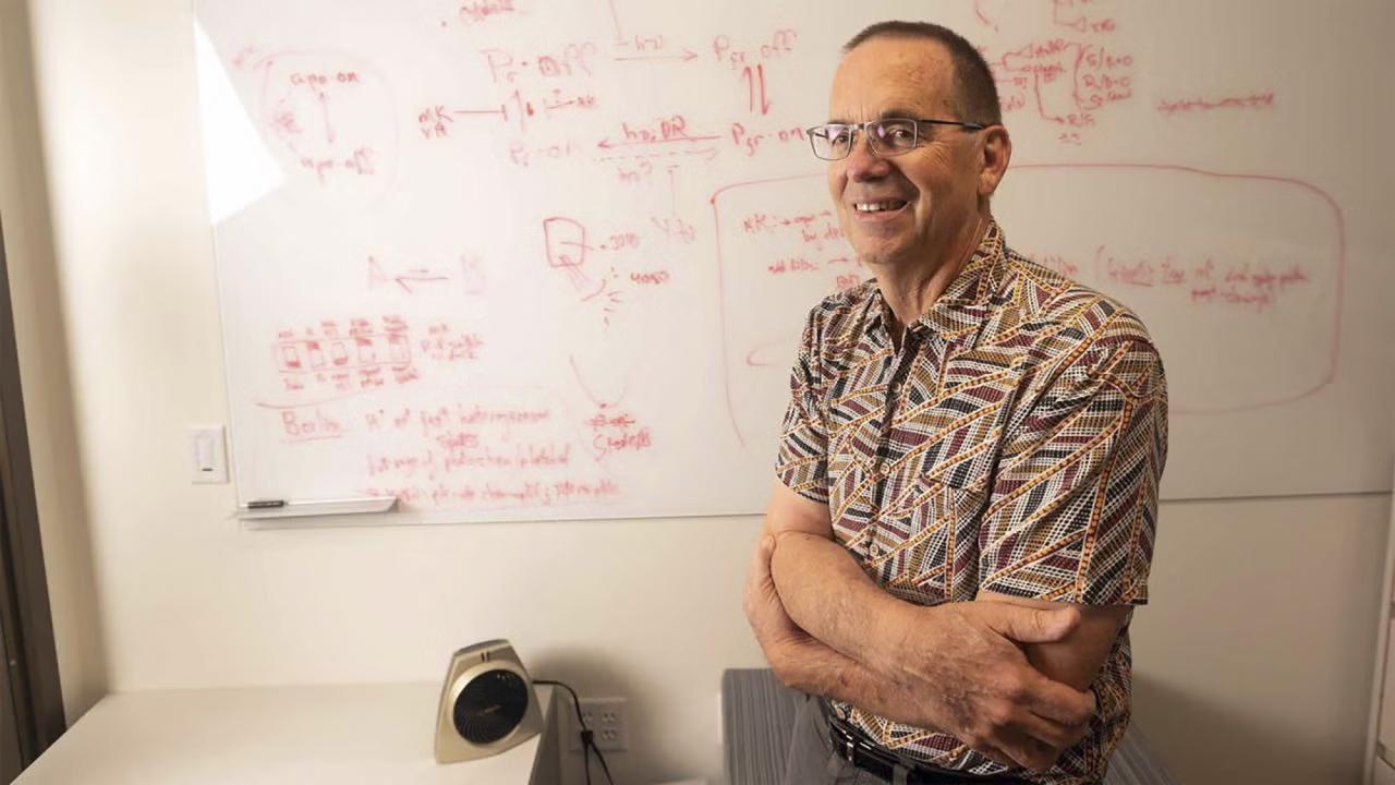 A man stands next to a whiteboard covered in scientific notes and diagrams, wearing glasses and smiling. He appears engaged and focused, conveying an atmosphere of learning and exploration.