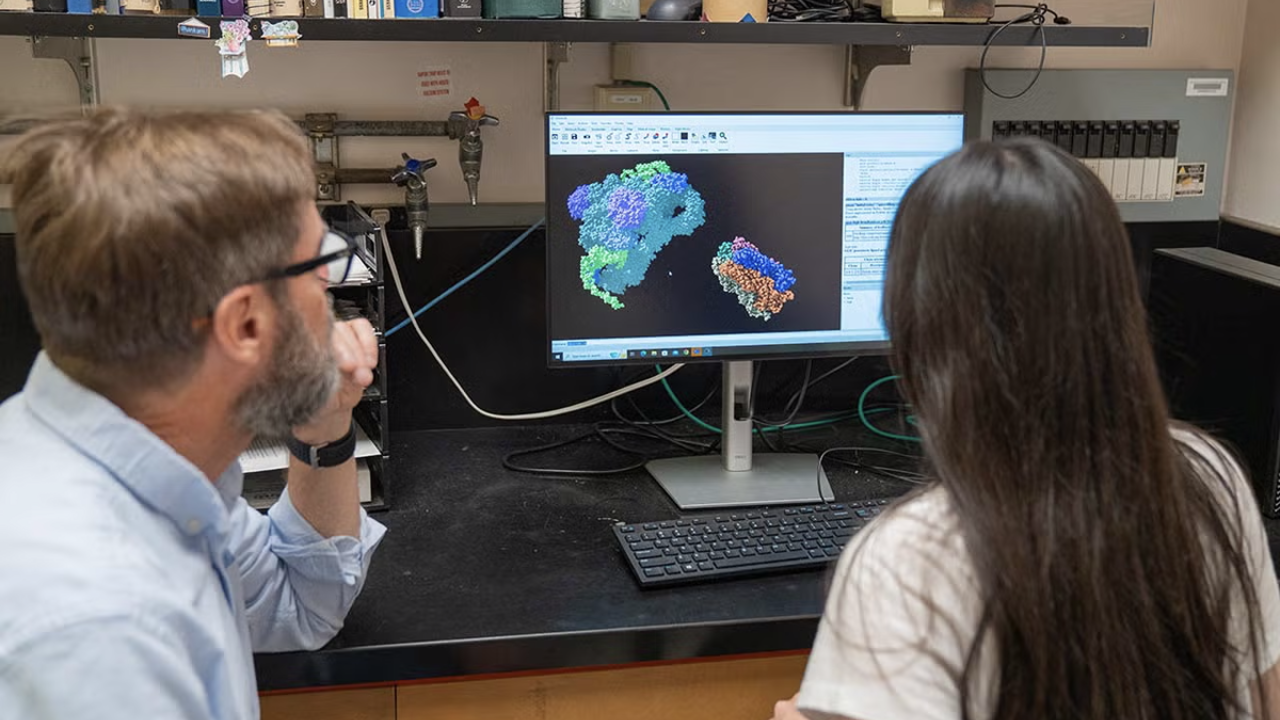 Two people sit at a lab bench, closely examining a colorful 3D molecular model displayed on a computer screen. The lab setting includes books, lab equipment, and coiled cables, indicating a research environment focused on molecular or structural biology.