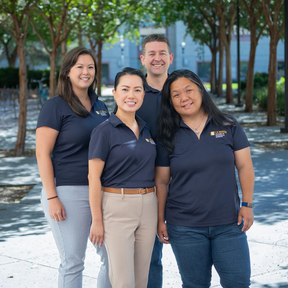Four UC Davis College of Biological Sciences staff in navy polos stand outdoors, smiling together under a row of shaded trees.