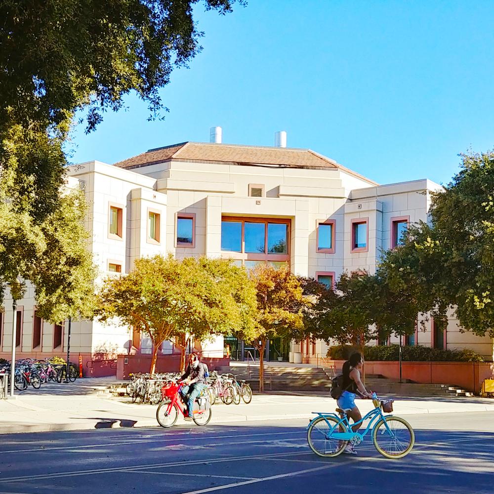 Students ride bikes across a sunny UC Davis campus in front of a white building framed by trees and bike racks.