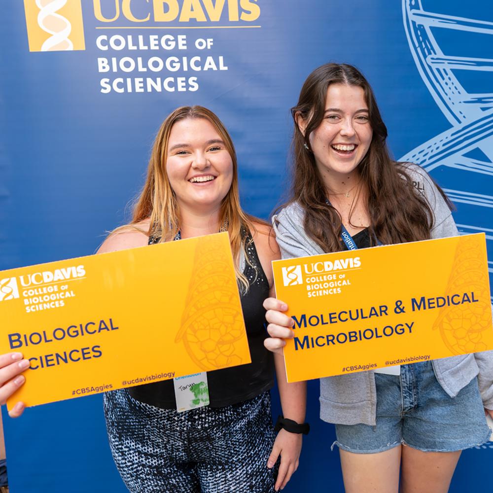 Two individuals smiling holding yellow signs in front of a UC Davis Backdrop