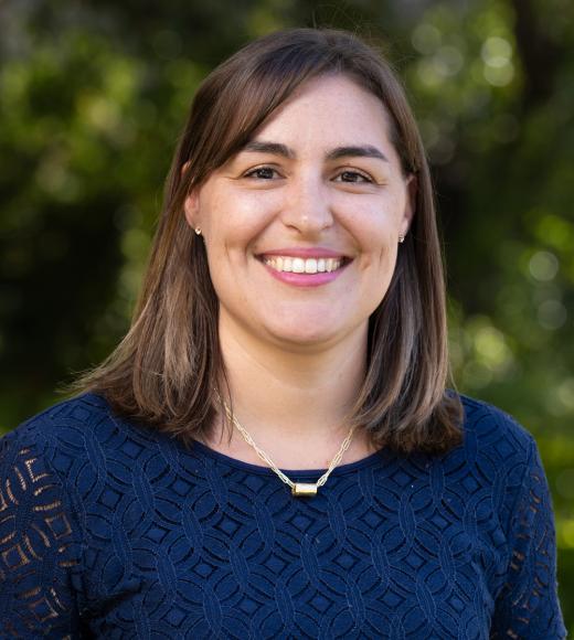 Portrait of a smiling woman with shoulder-length brown hair, wearing a navy lace-patterned top and a gold necklace, with a blurred green outdoor background.