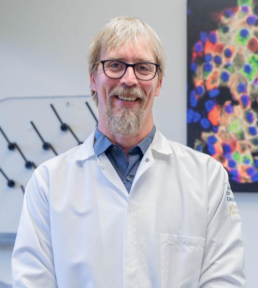 Smiling man with glasses and beard wearing a white lab coat standing in a lab with colorful cells image in background