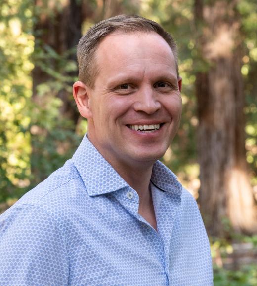 A smiling individual outdoors with a backdrop of lush greenery and tall trees, bathed in soft natural light, creating a warm and welcoming atmosphere.