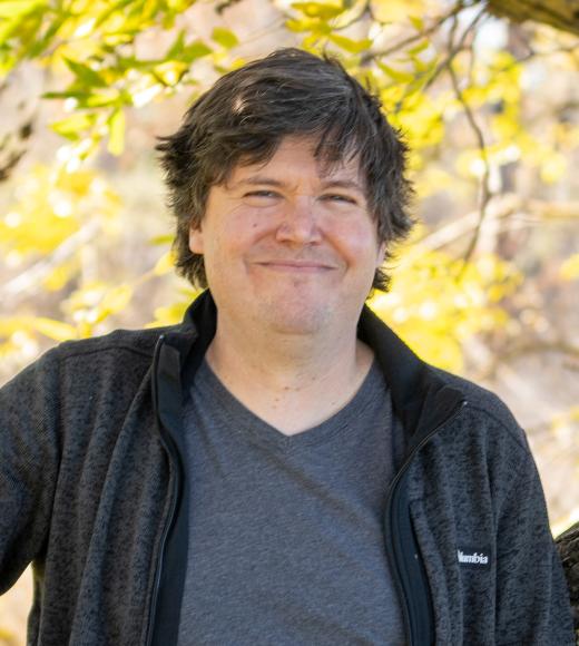 A person with short, slightly tousled hair wearing a gray shirt and dark jacket smiles outdoors in front of a background of yellow autumn leaves.