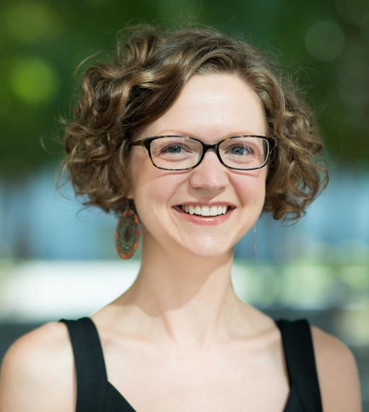 Portrait of a smiling individual with curly brown hair, wearing glasses, a black sleeveless top, and large earrings, set against a blurred outdoor background with greenery and soft lighting.