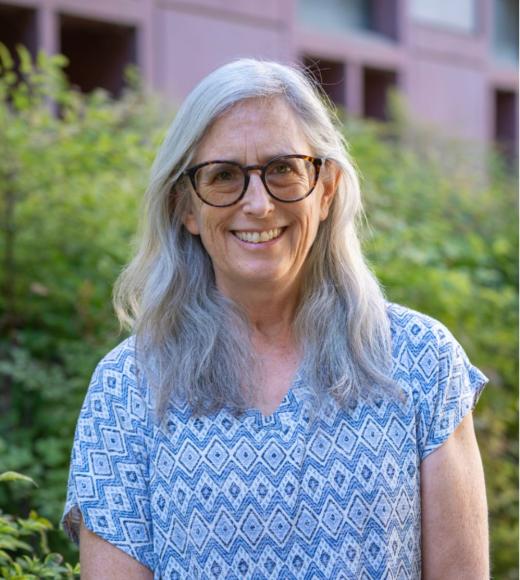 Person smiling outdoors wearing a blue patterned top, long gray hair and glasses, greenery behind.