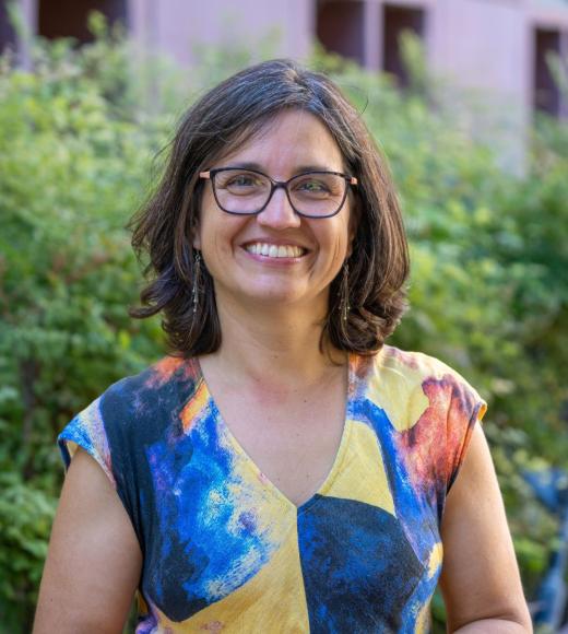 Person smiling outdoors in a colorful patterned top, glasses, and short dark hair, greenery behind.