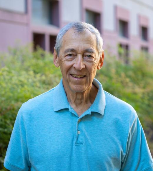 Person smiling outdoors in a light blue polo shirt, gray hair, greenery and building background.