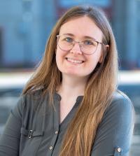 Woman with long hair and glasses smiles outdoors, wearing a dark button-up shirt with arms folded.