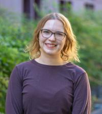 Smiling person with glasses and shoulder-length hair wearing a dark top, standing outdoors with greenery in the background.