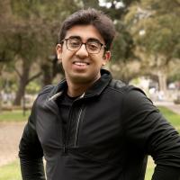 Kush Narang, a student with short dark hair and glasses, wearing a black zip-up jacket, smiles while standing outdoors with trees and a pathway in the background.