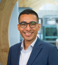 A smiling man wearing glasses and a navy blue suit jacket over a white dress shirt, standing in a modern building with curved wooden architecture in the background.