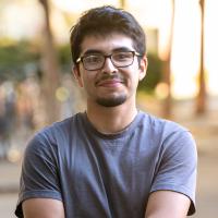 A young man with short dark hair, glasses, and a neatly trimmed beard smiles warmly at the camera, standing outdoors with his arms crossed. He is wearing a gray T-shirt, with blurred trees and sunlight in the background.