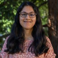A UC Davis student smiles softly while standing outdoors in front of leafy green trees. She has long dark hair, wears glasses, and is dressed in a peach-colored floral top.