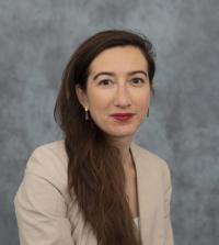 A person with long straight hair in a tan blazer in front of a grey background