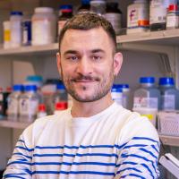 Person in a striped shirt stands smiling with arms crossed in a lab, shelves of chemical bottles visible behind.