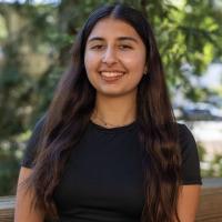 A UC Davis student smiles while standing outdoors in front of a background of trees. She has long dark hair, wears a black top, and a delicate beaded necklace.