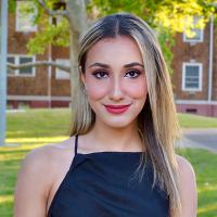 Smiling young woman in a black dress standing outdoors, with trees and a brick building in the background.