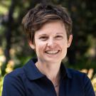 A woman with short brown hair and a friendly smile wears a navy UC Davis College of Biological Sciences polo shirt, standing outdoors with a blurred green background.