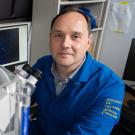 A scientist in a blue University of California lab coat sits beside a microscope and imaging equipment in a research lab. A computer screen behind him displays multicolored microscopic images.