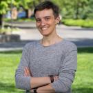 A person with short hair and a gray long-sleeve shirt stands smiling with arms crossed on a sunny, tree-lined lawn at UC Davis.