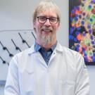 Smiling man with glasses and beard wearing a white lab coat standing in a lab with colorful cells image in background