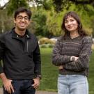 Two students standing outdoors on a green lawn, smiling at the camera. The student on the left wears glasses and a black jacket with hands in pockets, while the student on the right wears a multicolored sweater and light jeans with arms crossed. Trees and pathways are visible in the background.