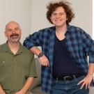 Two people smile in a bright room one seated on a couch and the other standing with an arm resting on the couch back.