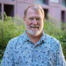 Person smiling outdoors, wearing a blue patterned button-up shirt with greenery and a building behind.