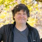 A person with short, slightly tousled hair wearing a gray shirt and dark jacket smiles outdoors in front of a background of yellow autumn leaves.