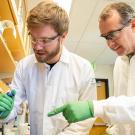 Two researchers in lab coats and standing in a laboratory setting examining an object on a table.
