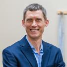 Portrait of a middle-aged man with short, graying hair, wearing a navy blue blazer over a light blue dress shirt. He is smiling warmly and looking directly at the camera, with a neutral beige background featuring a blurred object on the right side.